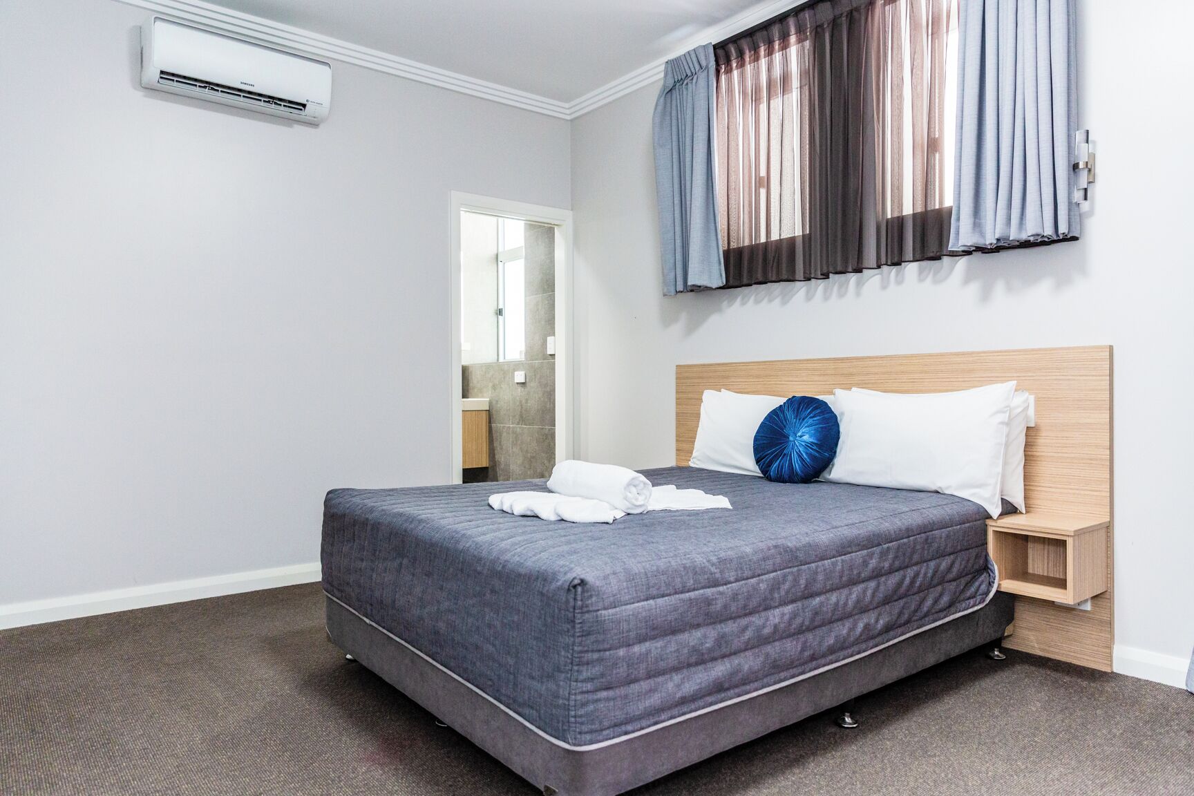 A hotel room with a large bed, a wooden headboard, a nightstand, and a wall-mounted air conditioner.