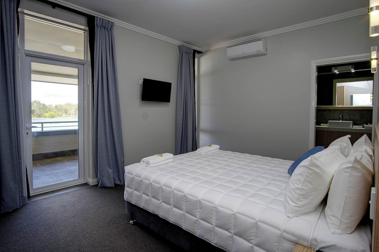 Hotelkamer met een bed, een flatscreen-tv, een raam met gordijnen, een airconditioning en een badkamer met een wastafel en spiegel.