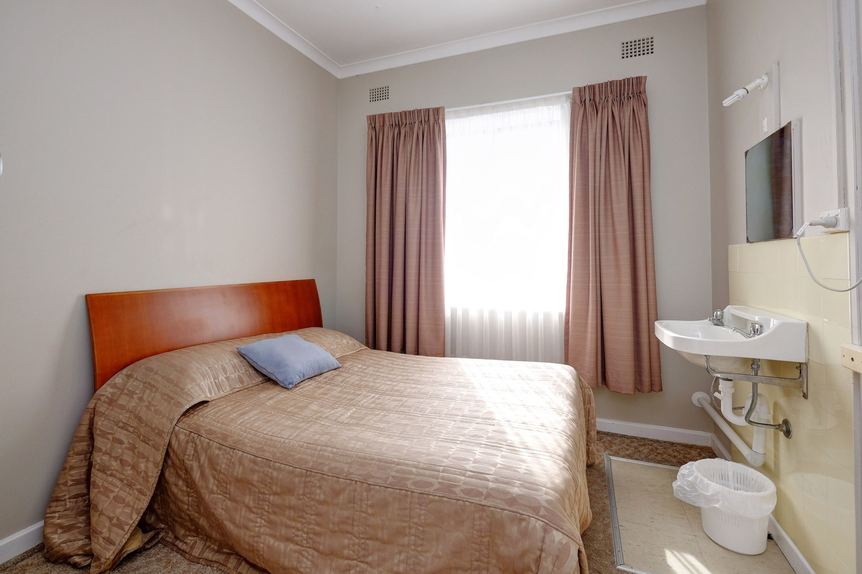A hotel room with a bed, a single pillow, a window with curtains, a sink, and a trash bin.