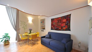 Un salón con sofá azul, sillas amarillas, una mesa de comedor con un jarrón, un tapiz en la pared y un teléfono.