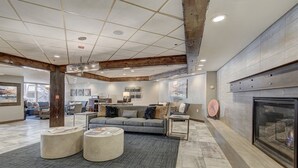 A spacious lobby with a grey sofa, two round coffee tables, a fireplace, and a high ceiling with exposed beams. - Village Majesty (Breckenridge)