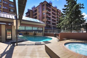 A swimming pool area with a hot tub, wooden deck, and a building with balconies in the background. - Liftside Studio (Breckenridge)