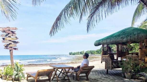 A beachside seating area with wooden tables and chairs, a thatched-roof structure, and a signpost with directions. - Coco Paradise Beach Resort (Currimao)