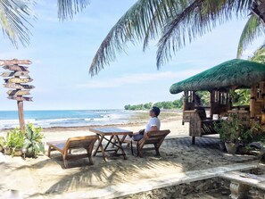 Ein Sitzbereich direkt am Strand mit Holztischen und -stühlen, einer Reetdachkonstruktion und einem Wegweiser mit Beschilderung.