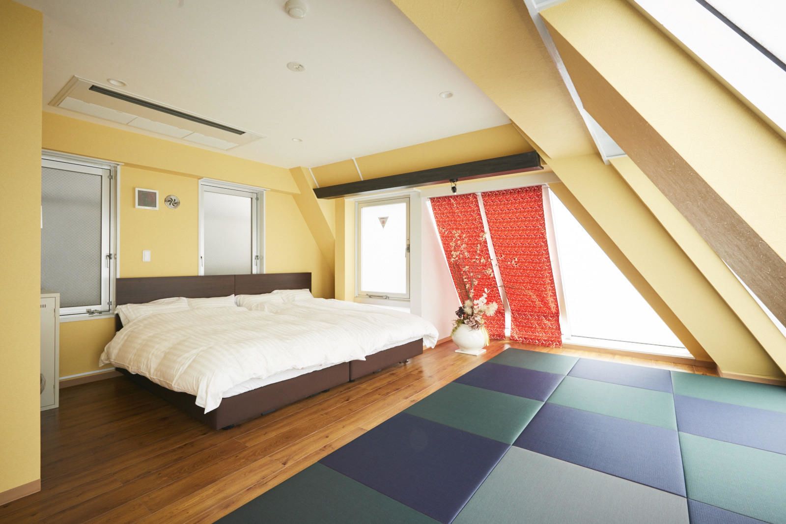 A bedroom with a large bed, wooden flooring, a window with a red curtain, and a skylight.