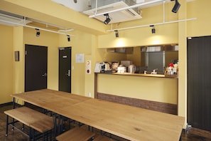Un salón de conferencias con una larga mesa de madera, varias sillas, una kitchenette con una máquina de café y un detector de incendios en la pared.