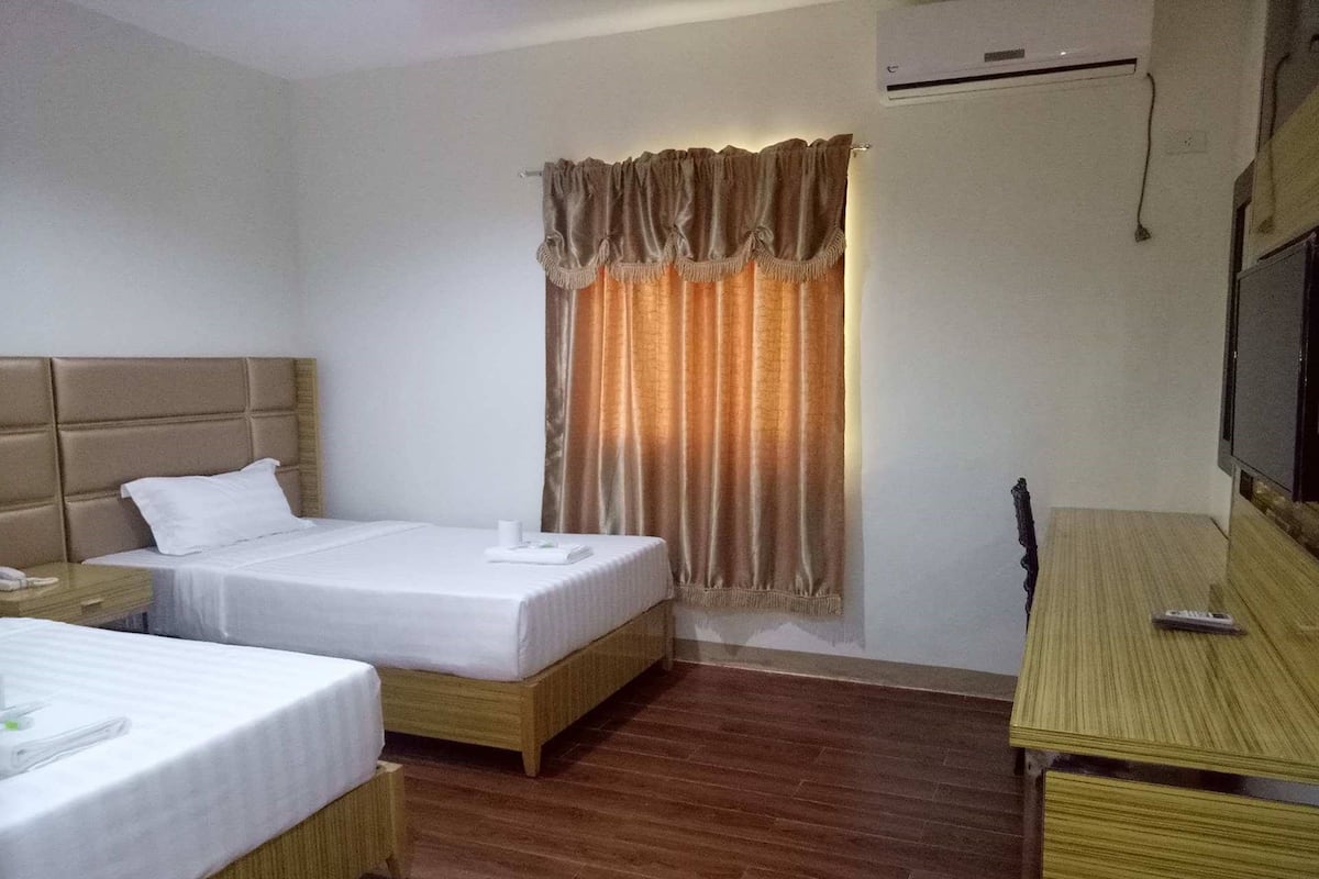 A hotel room with two single beds, a wooden headboard, a wooden desk, a television, and an air conditioning unit.