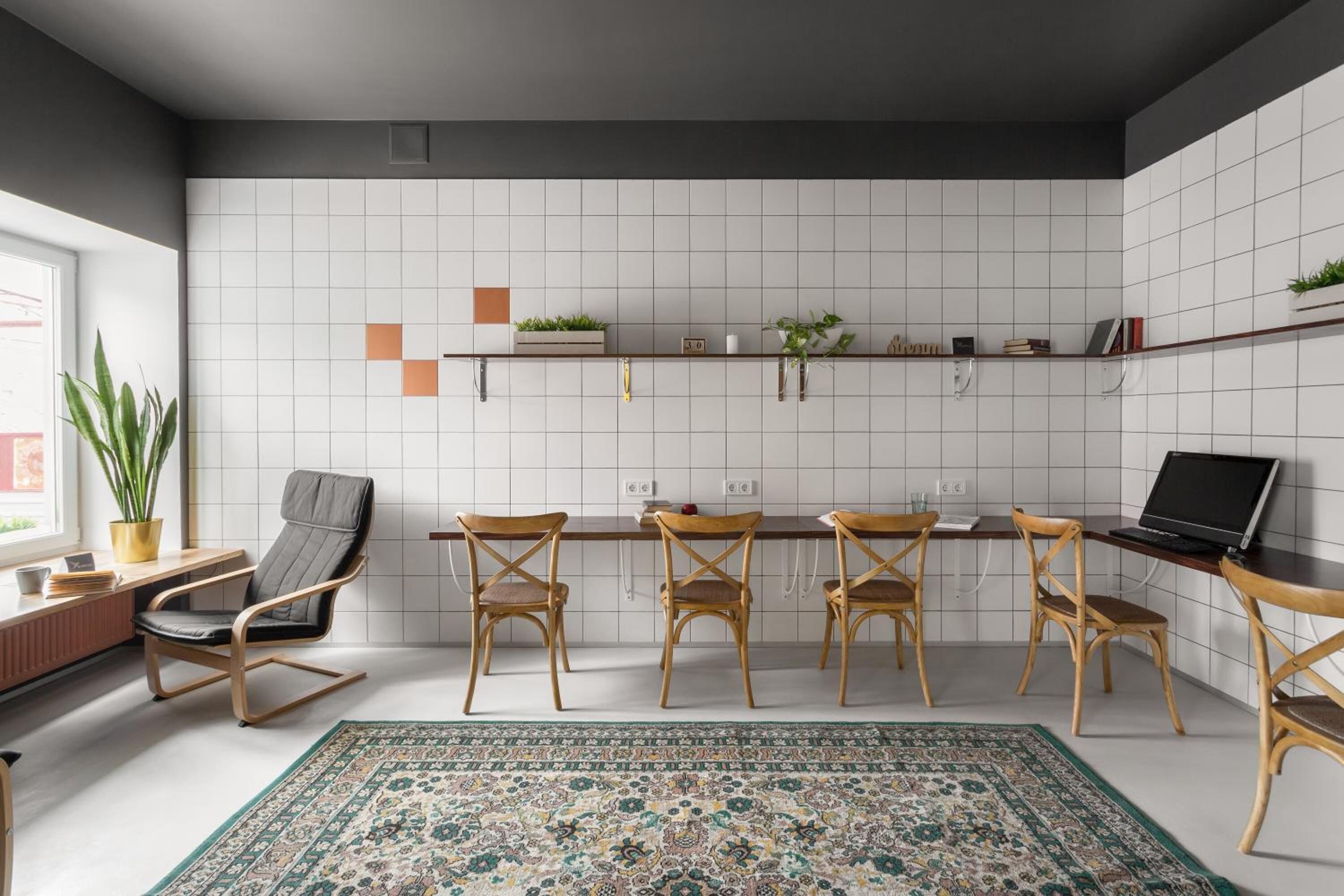 A modern kitchen with white tiles, wooden chairs, a dining table, and a laptop on a desk.