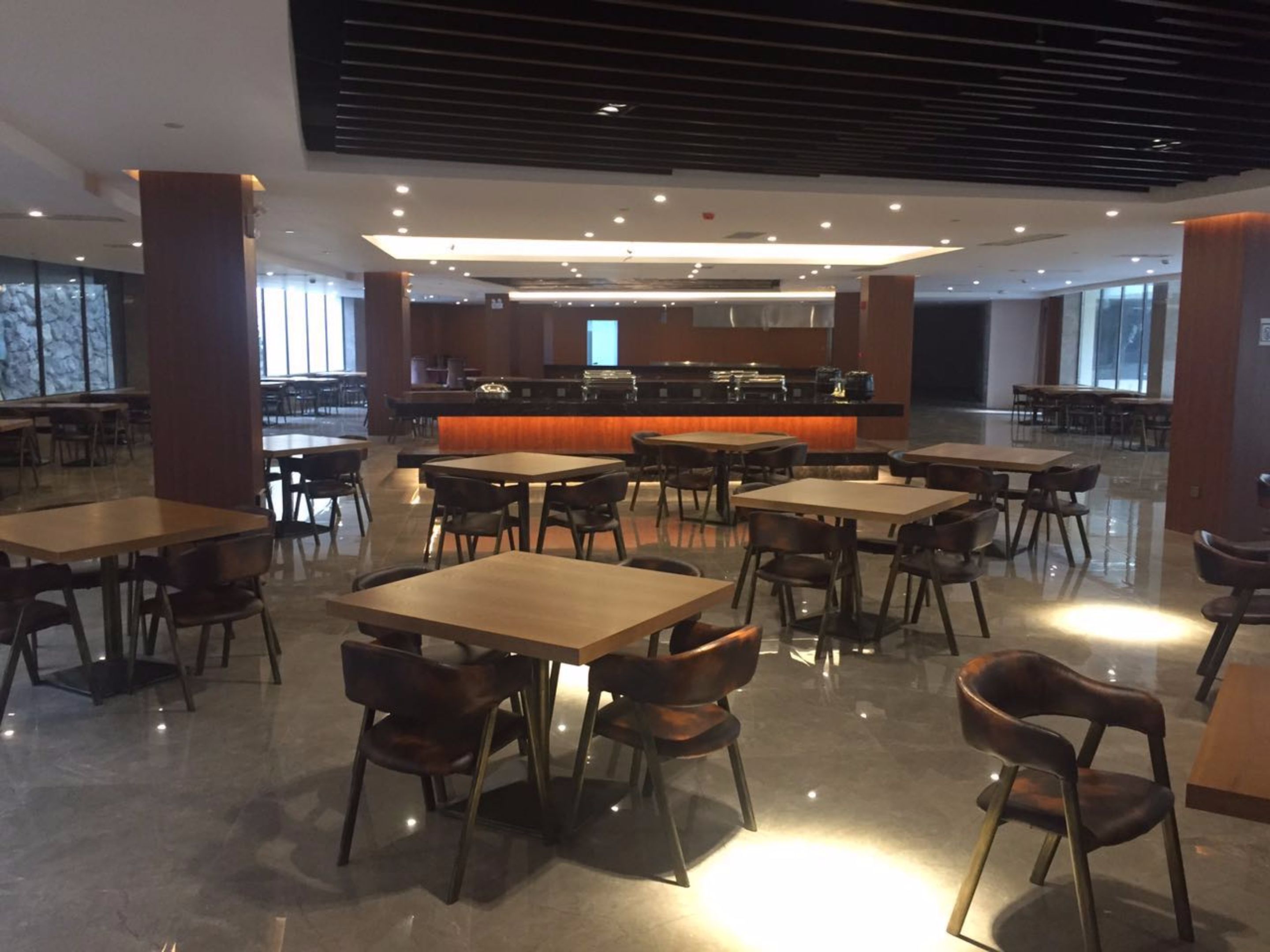 A modern dining area with multiple tables and chairs, a central bar area, and large windows providing natural light.