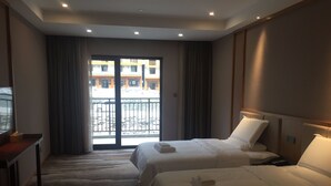 Een hotelkamer met twee bedden, een bureau en een groot raam met uitzicht op gebouwen.