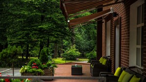 A covered outdoor seating area with wicker furniture and cushions, surrounded by lush greenery and a brick building.