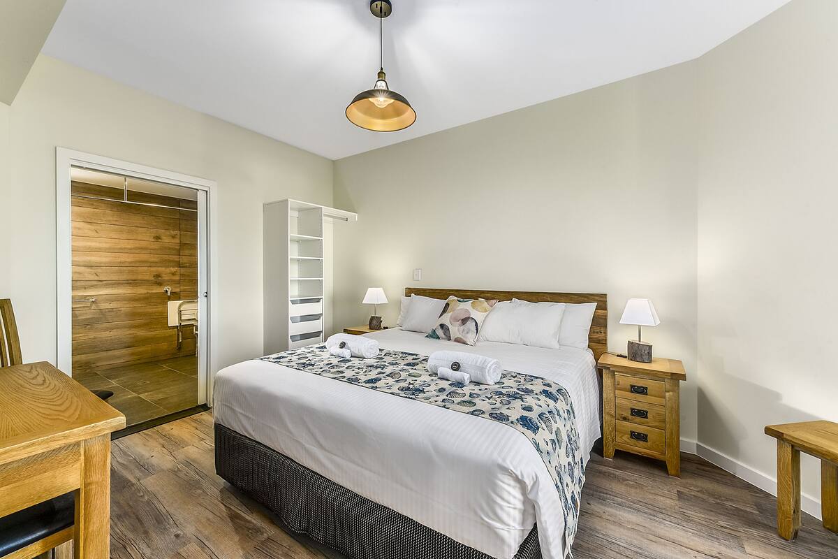 Une chambre avec un plancher en bois, un lit recouvert d’une couvre-lit à motifs floraux, deux tables de chevet avec des lampes, une armoire en bois et un petit banc en bois.