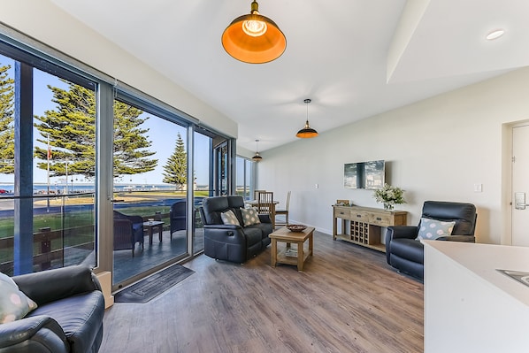 A spacious living room with a large glass door leading to a balcony, a wooden floor, and a modern light fixture.