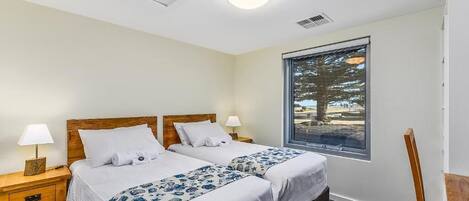 Habitación de hotel con dos camas, una ventana que da a árboles, una mesita de noche de madera con una lámpara y una silla.