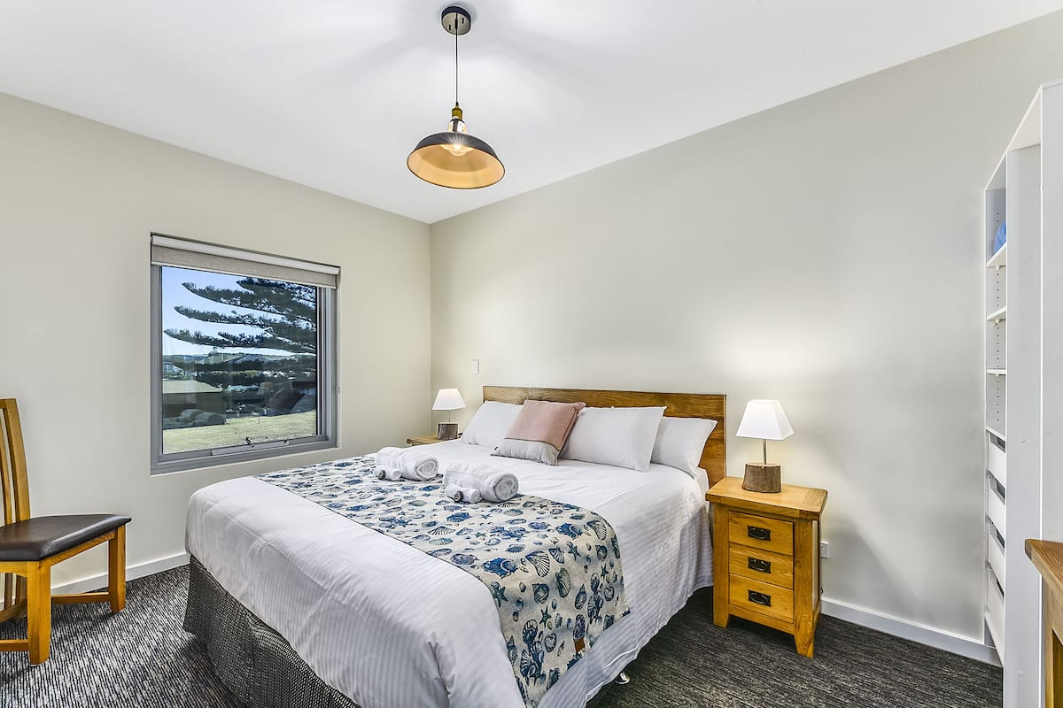 Une chambre d’hôtel bien aménagée, avec un grand lit, une fenêtre donnant sur des arbres, une table de chevet en bois avec une lampe et une chaise.