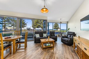 Amplia sala de estar con una mesa de comedor de madera, dos sofás de cuero negro, una mesa de centro y un armario de madera. Ventanales ofrecen vistas al exterior.