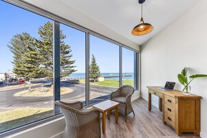 Una habitación con un gran ventanal que da a una carretera y árboles, una silla de mimbre, una mesita y un escritorio de madera con un ordenador portátil y una planta.