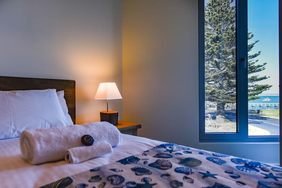 Un lit bien fait, avec une serviette enroulée et une lampe sur une table de chevet, une fenêtre donnant sur des arbres et une route.