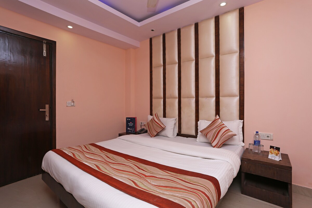 A hotel room with a large bed, a wooden headboard, a nightstand with a water bottle and snacks, and a door with a handle.