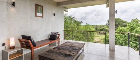 Un balcón amplio con banca de madera y una mesa de centro, pared de hormigón con un cuadro enmarcado, y vistas a zonas verdes.
