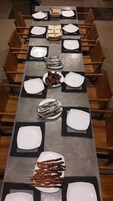 Una larga mesa de comedor dispuesta con platos, sándwiches, alimentos envueltos en papel de aluminio y brochetas, rodeada de sillas de madera.