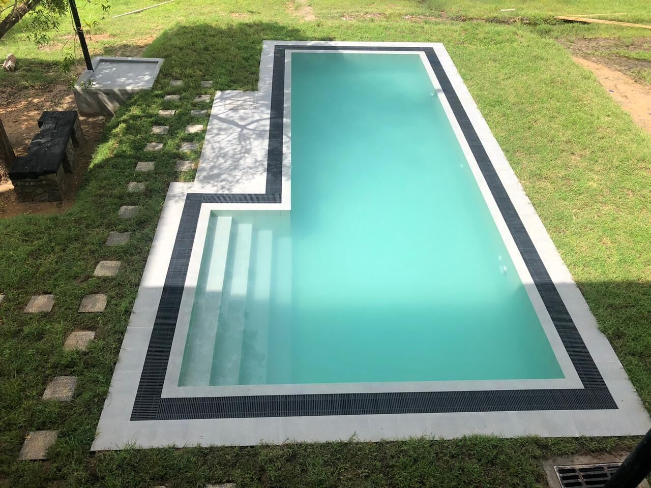 A rectangular swimming pool with a tiled edge, surrounded by a grassy area with stepping stones leading to a covered structure.