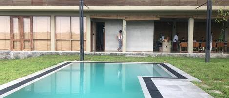 A swimming pool with a tiled deck, a building with a flat roof and large windows, and a grassy area.
