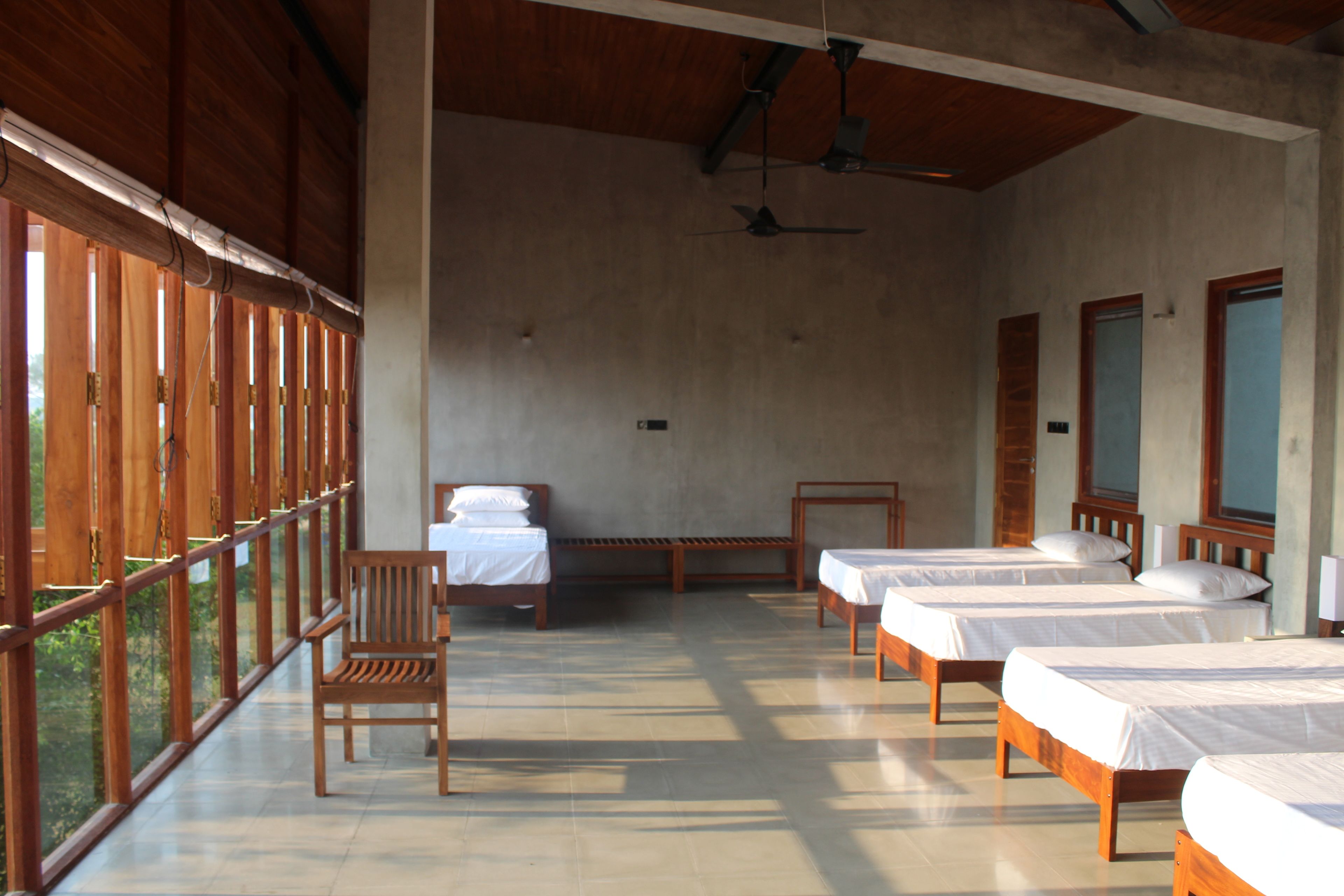 A room with four beds aligned along the wall, a wooden chair, and a ceiling fan.