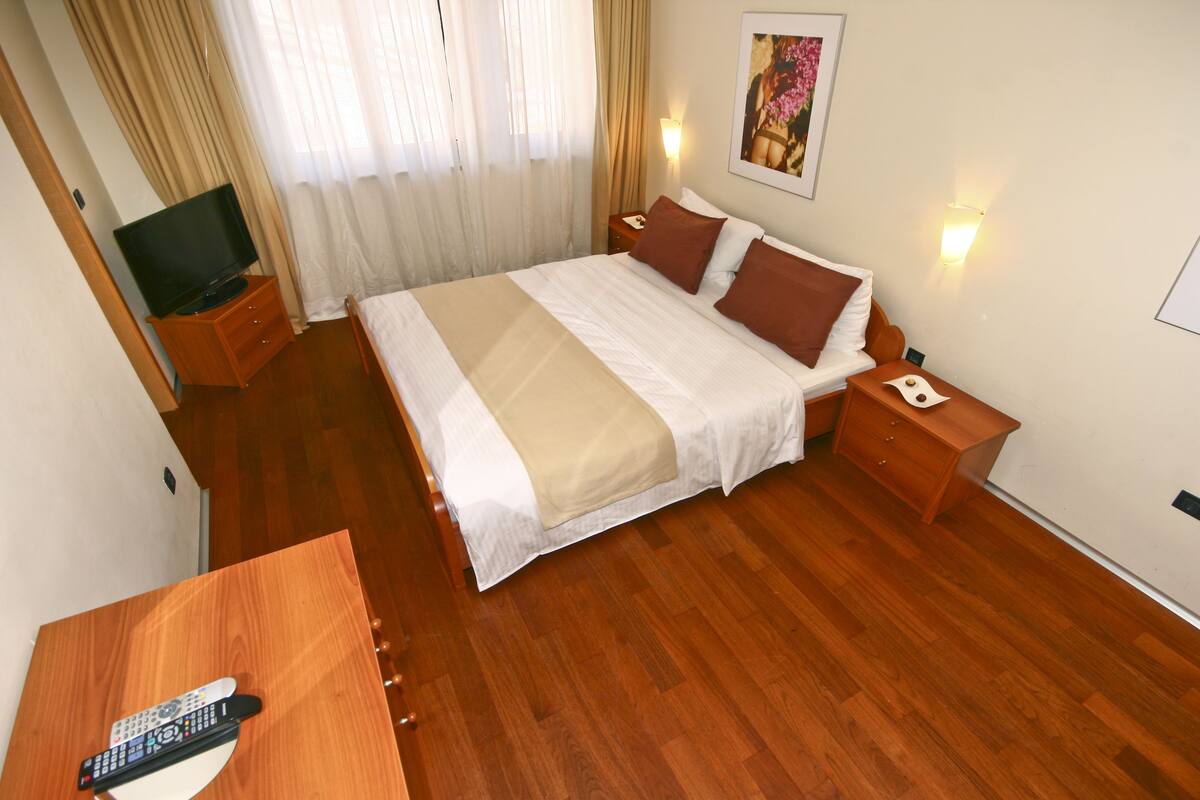 A hotel room with a bed, wooden nightstands, a TV, and a remote control on a desk.