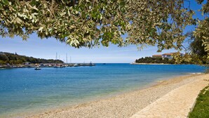 A pebble beach with boats in the water, a clear sky, and buildings in the background. - Villa Feniks (Medulin)