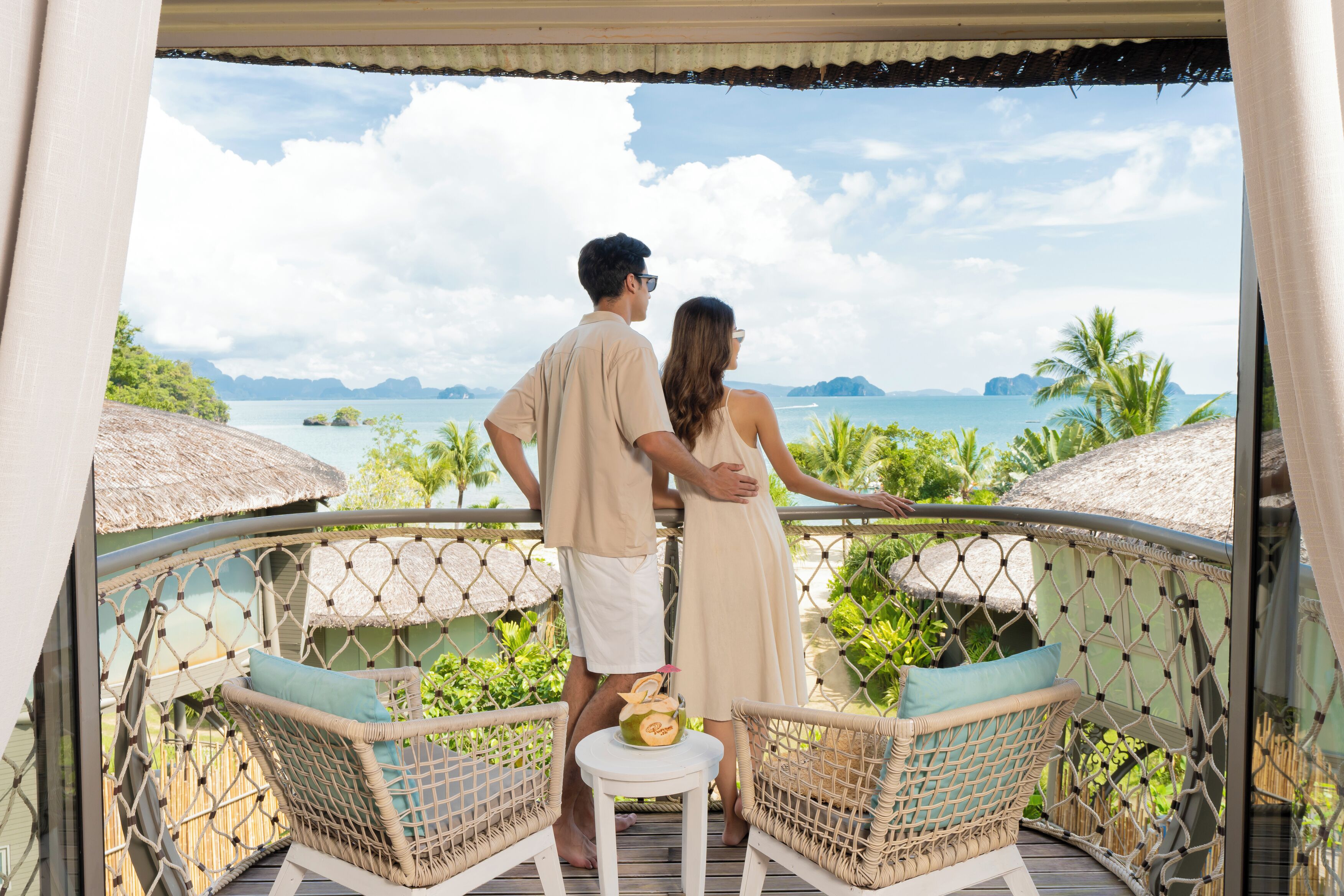 A couple standing on a balcony overlooking a tropical beach with thatched roofs and lush greenery.