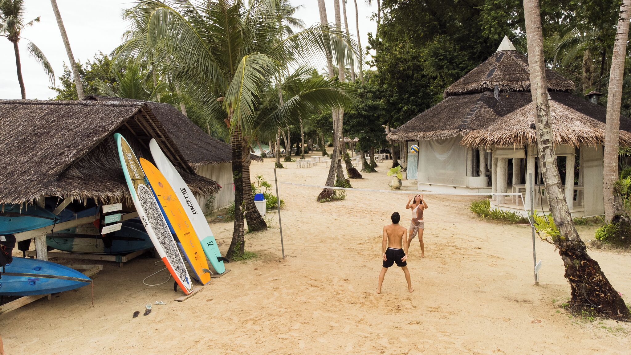 Ein Strand mit strohgedeckten Hütten, einem Volleyballnetz und Surfbrettern, die an einer Hütte lehnen.