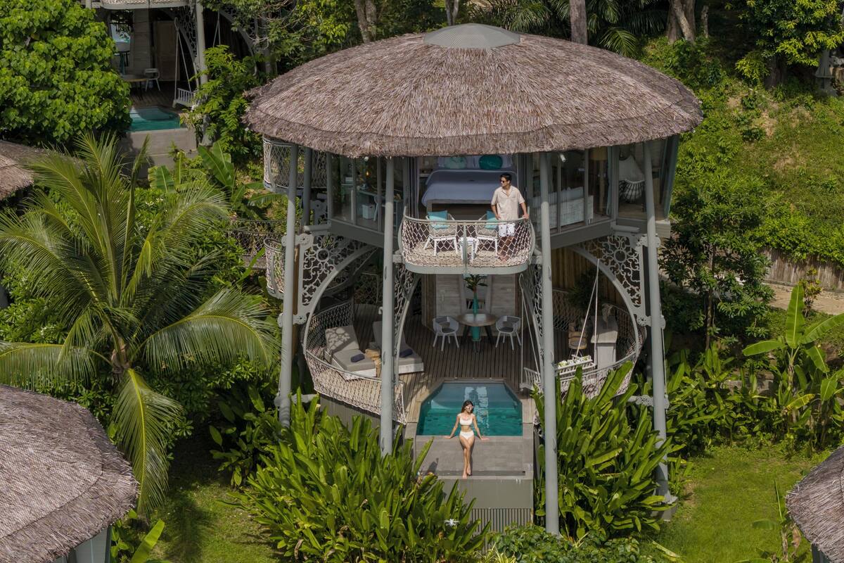 A multi-level outdoor pool area with a thatched roof structure, surrounded by lush greenery and palm trees.