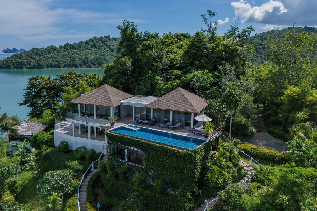 A modern villa with a swimming pool, surrounded by lush greenery and overlooking a large body of water.