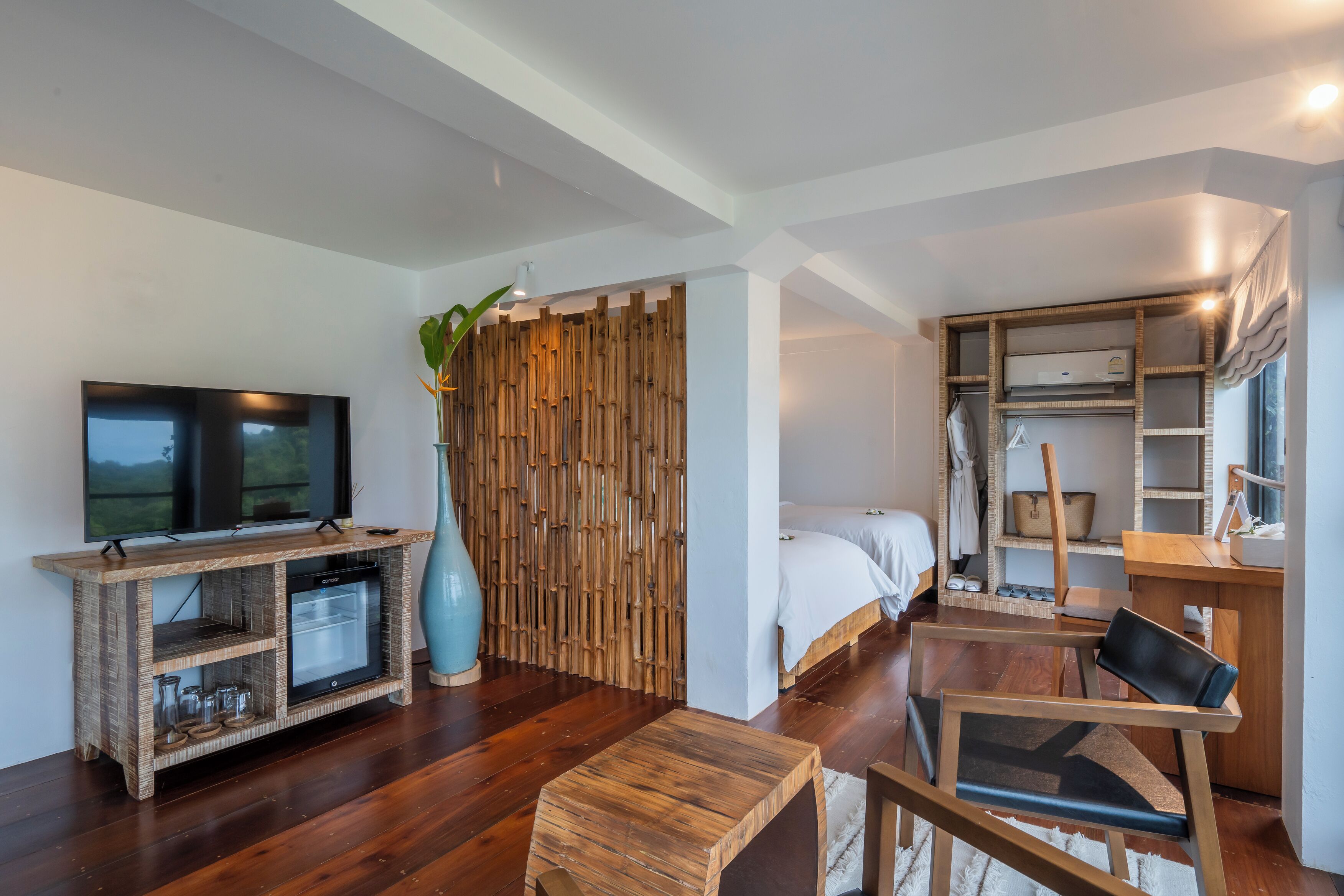 A modern room with a flat-screen TV, a wooden console table, a small refrigerator, a large vase, a bed with white bedding, a wooden chair, and a built-in wardrobe.