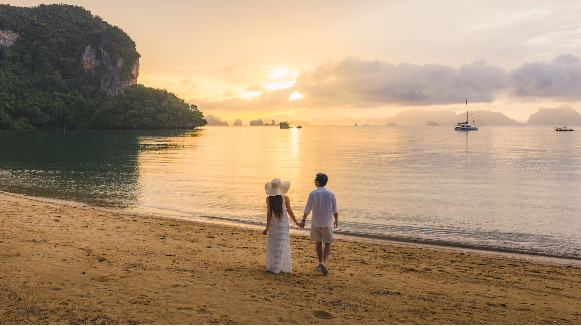 A beach with a couple holding hands, a boat, and a sunset.