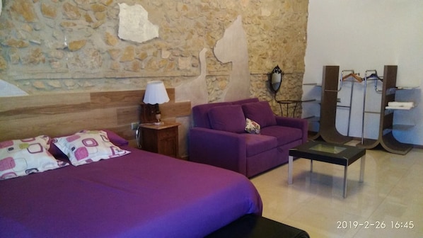 A room with a bed, a purple sofa, a small table, and a stone wall.