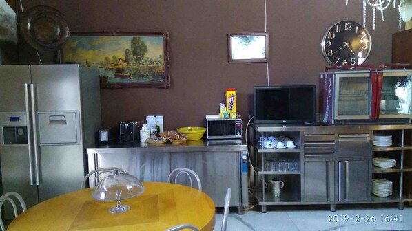 A compact kitchen with a refrigerator, microwave, and a small dining table with chairs. A television is mounted on the wall, and there are cabinets below it.