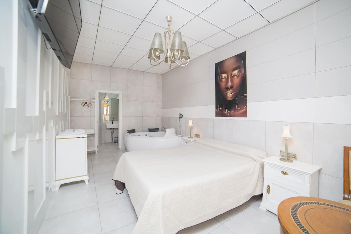 A modern hotel room with a large bed, a sitting area with a small table, a bathtub, and a large mirror.