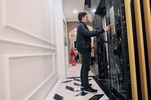 Una persona interactuando con la puerta de un ascensor de vidrio en un pasillo con paredes blancas y un piso de baldosas blanco y negro.