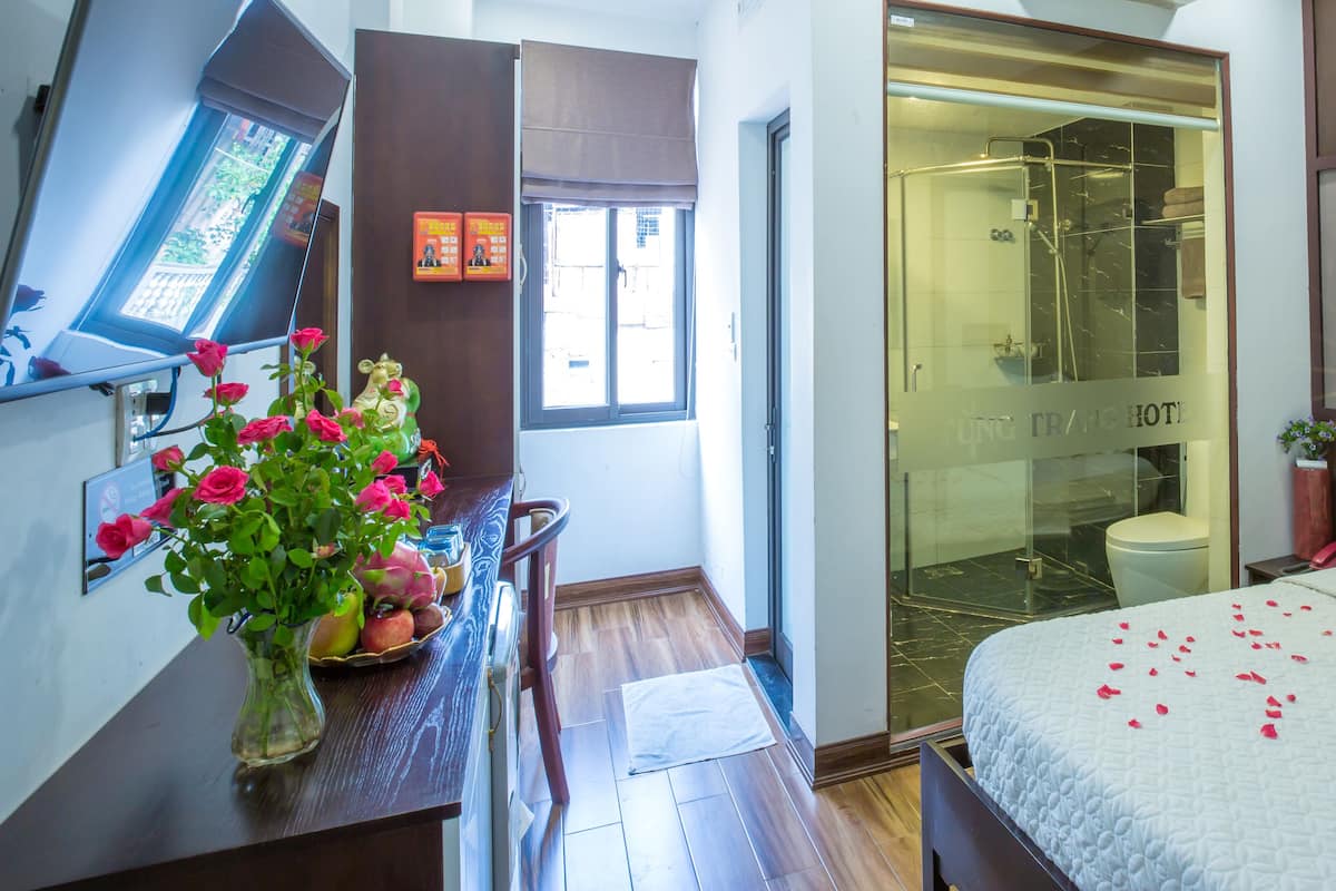 A hotel room with a bed, a desk with a vase of flowers, a bathroom with a glass shower, and a window with a view.