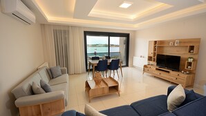 A modern living room with a sofa, a coffee table, and a TV stand. There is a large window with a view of the sea.