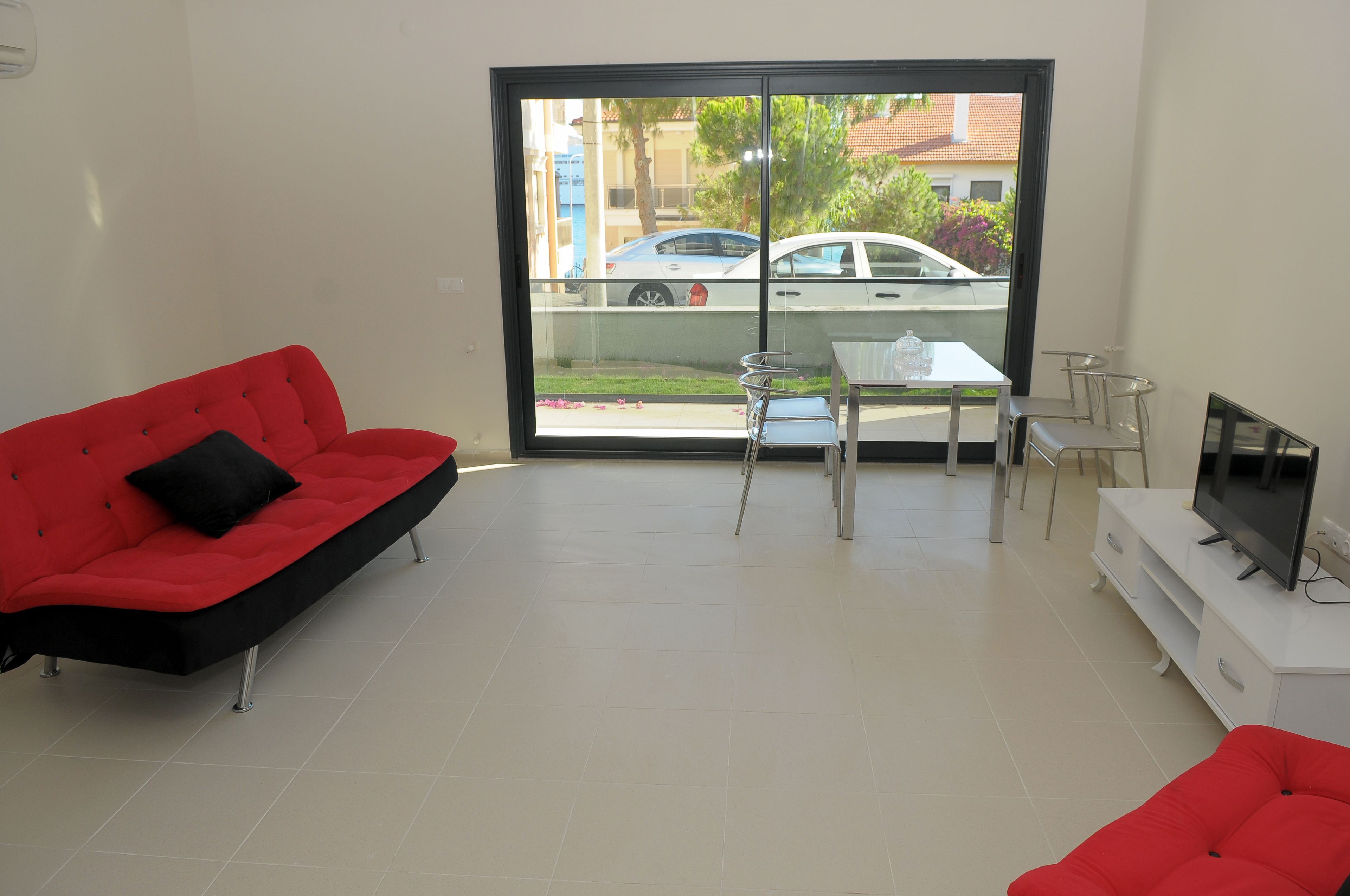 A modern living room with a red sofa, a glass door leading to a balcony, a small table, and a TV on a white stand.
