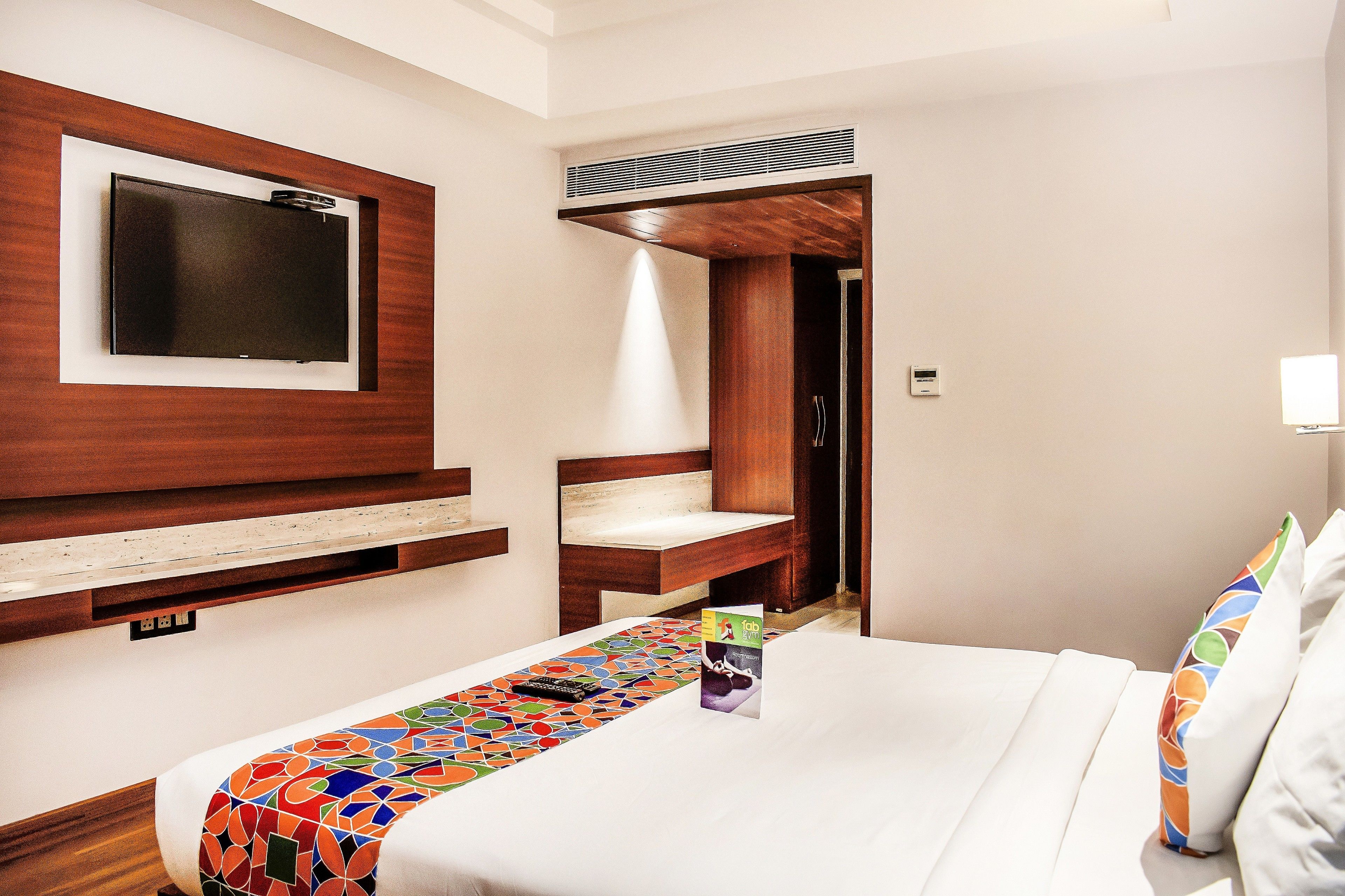 A hotel room with a bed, a TV mounted on the wall, a wooden headboard, a bedside table, and a small shelf.
