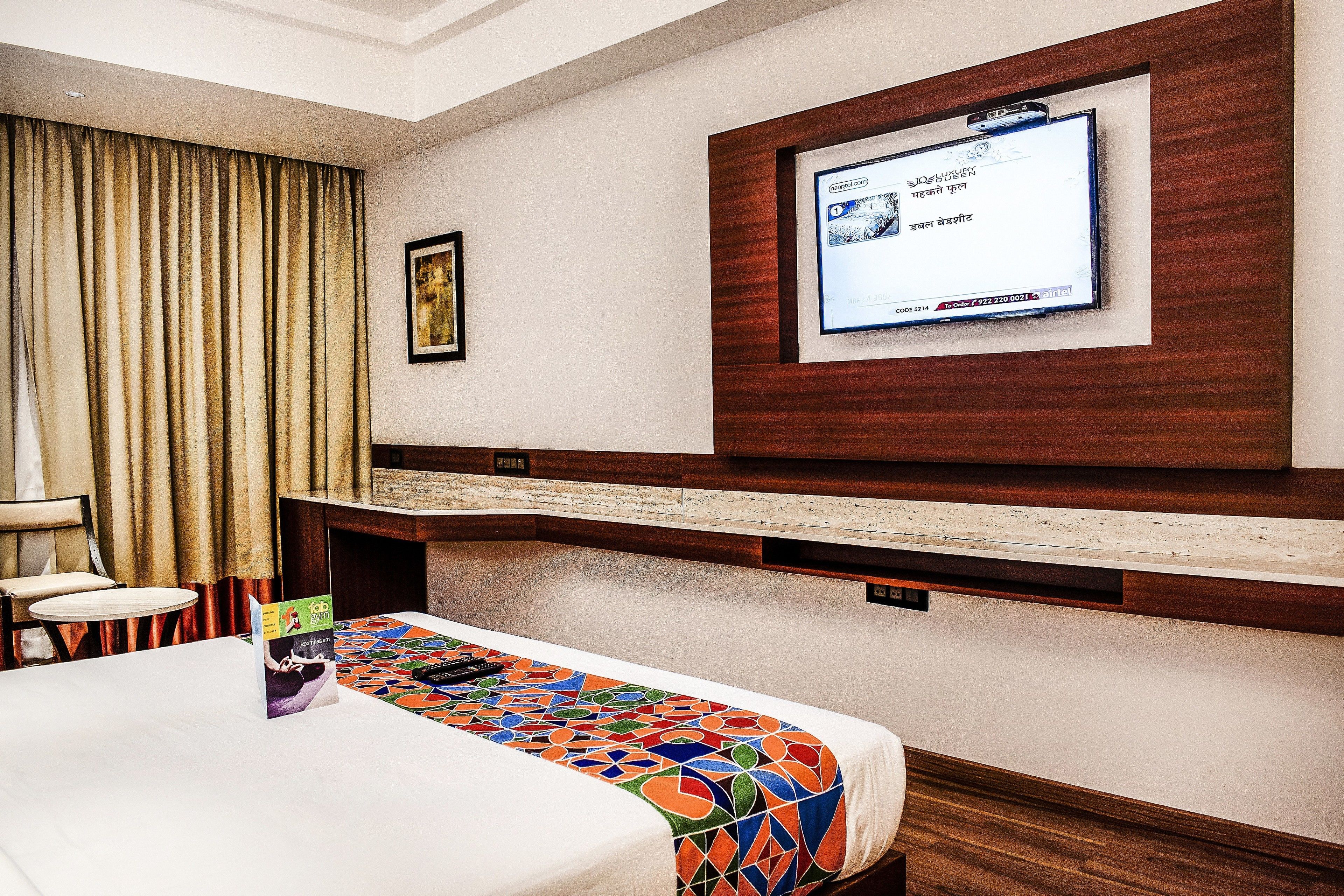 A hotel room with a bed, a desk, a chair, a TV mounted on the wall, and a window with curtains.