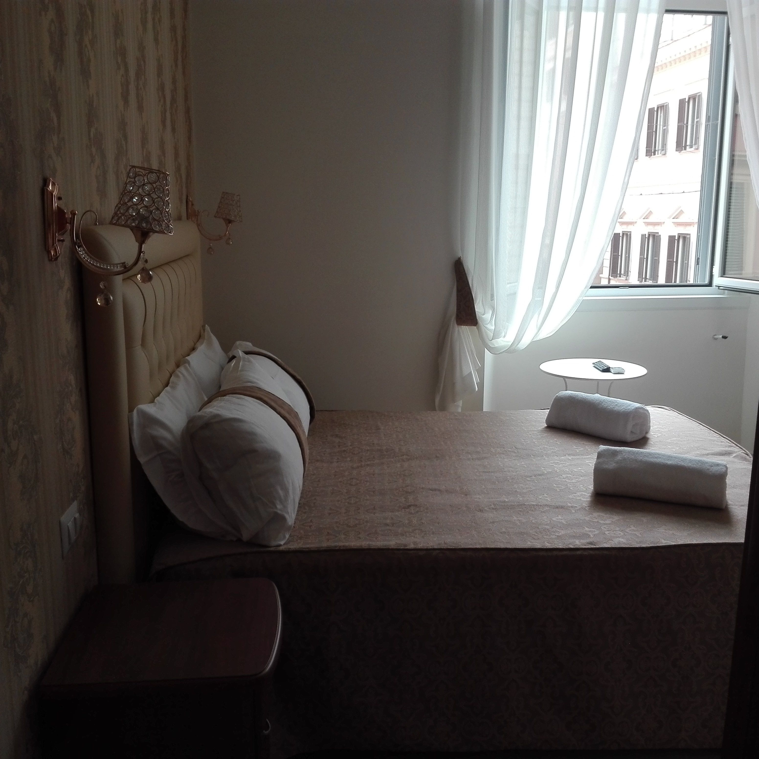 A hotel room with a bed, a small bedside table, a window with curtains, and a view of buildings outside.
