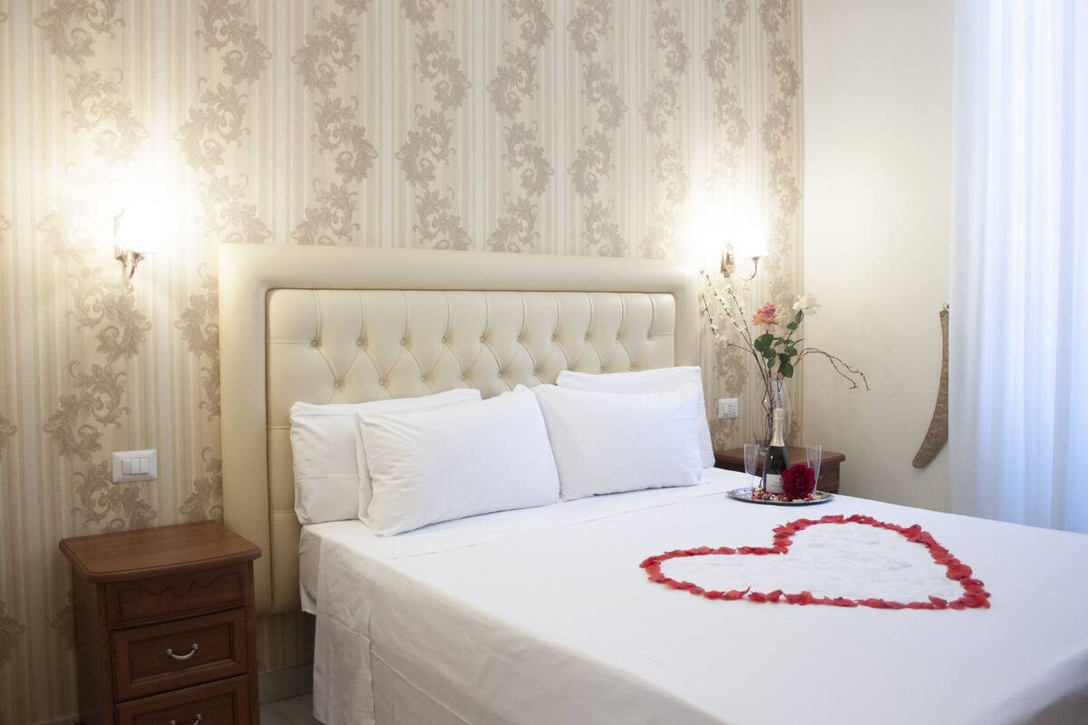 Una cama bien hecha con un adorno con forma de corazón en la colcha, una mesita de noche y un jarrón con flores.