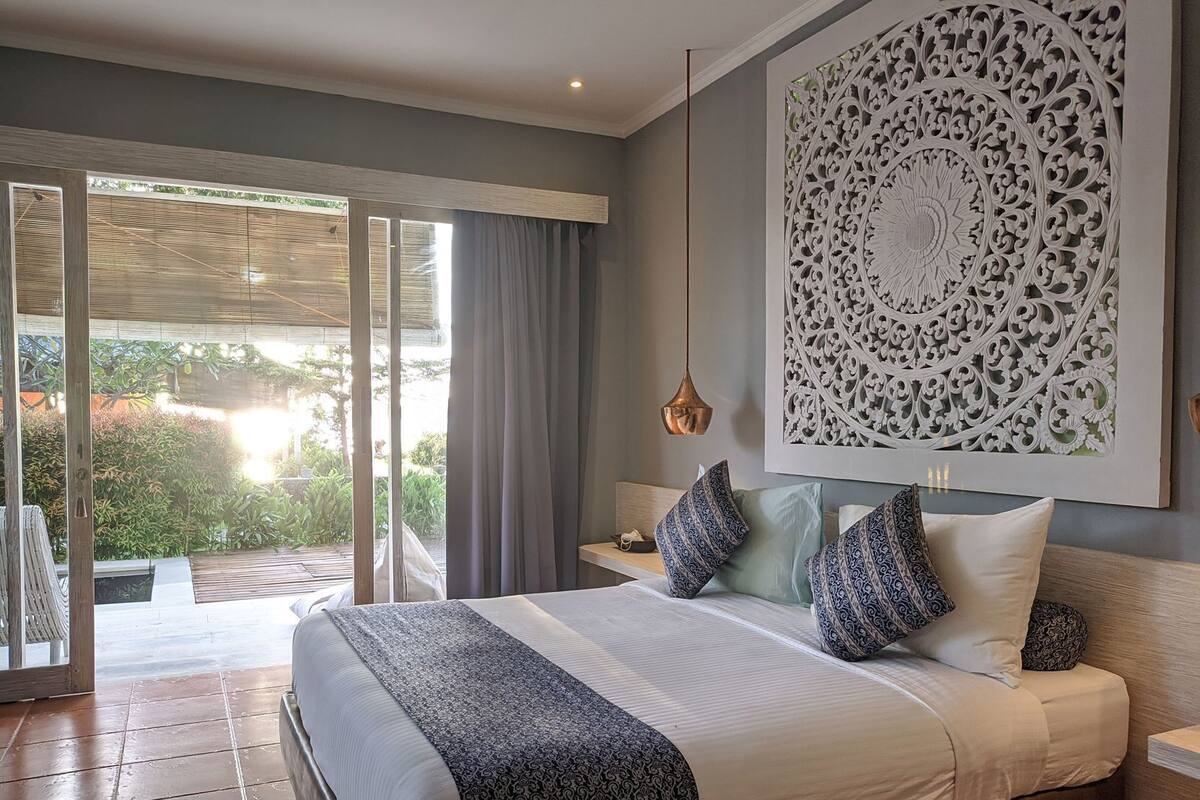 A bedroom with a large bed, a view of the outdoors, a decorative wall piece, and a hanging light fixture.