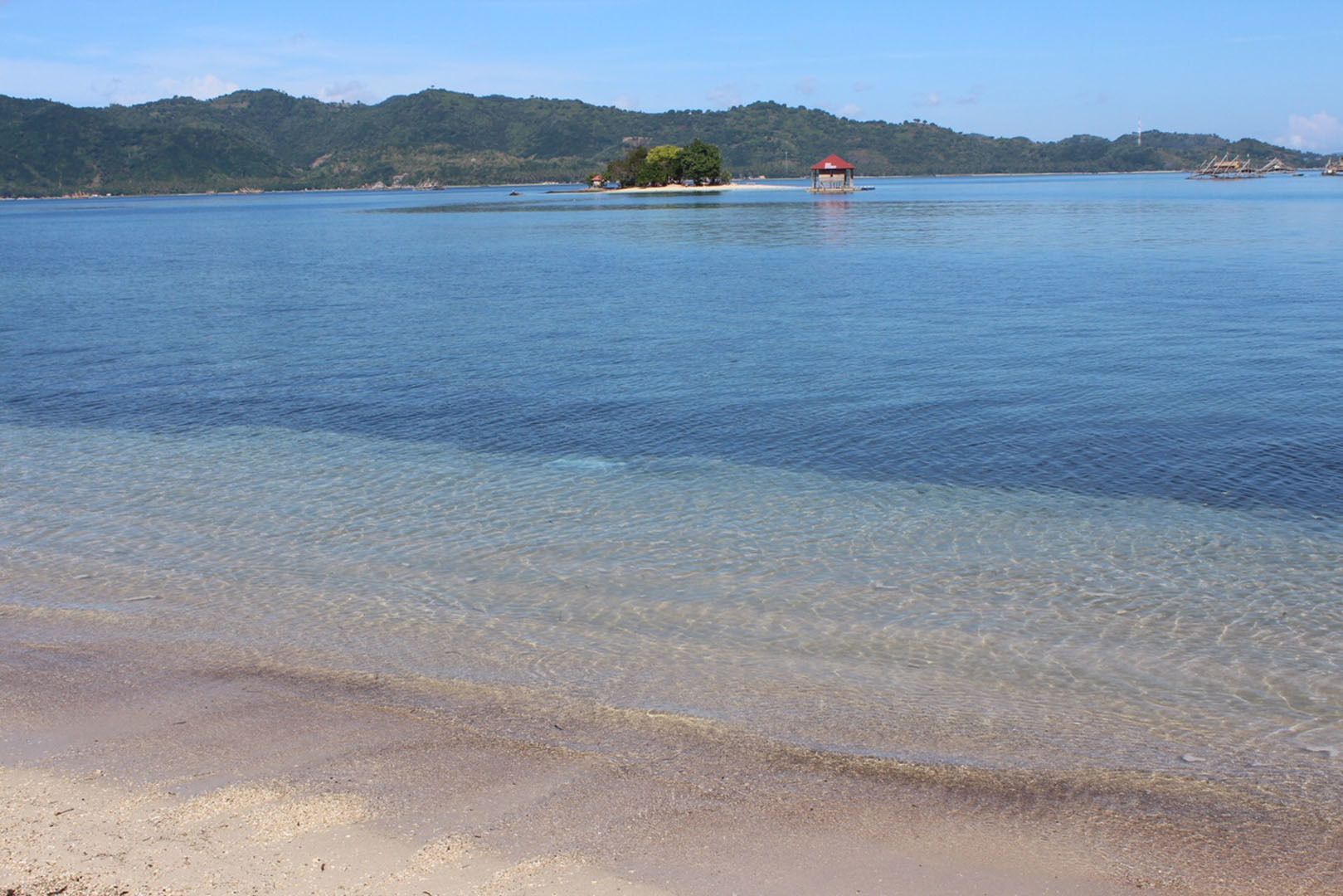 A beach with clear water, a small island with a red-roofed structure, and a distant hill.