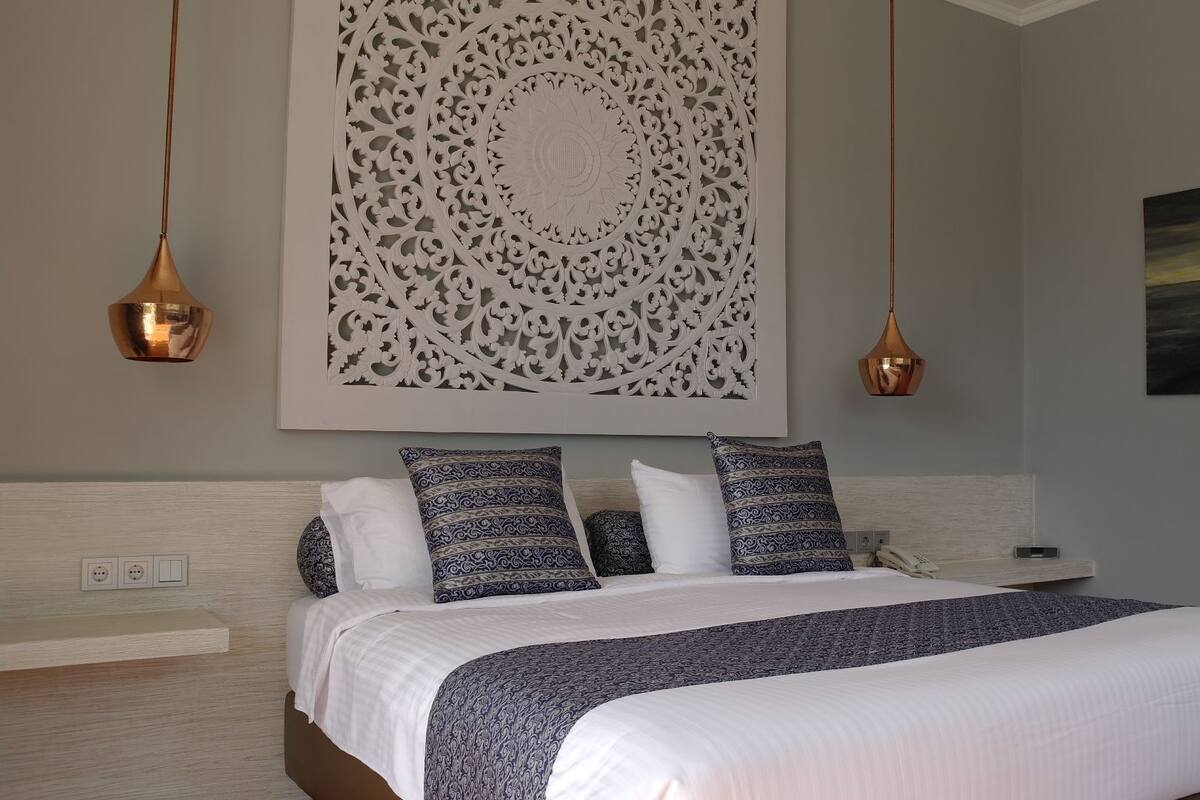 A bedroom with a large bed, patterned pillows, a decorative wall panel, and two pendant lights.